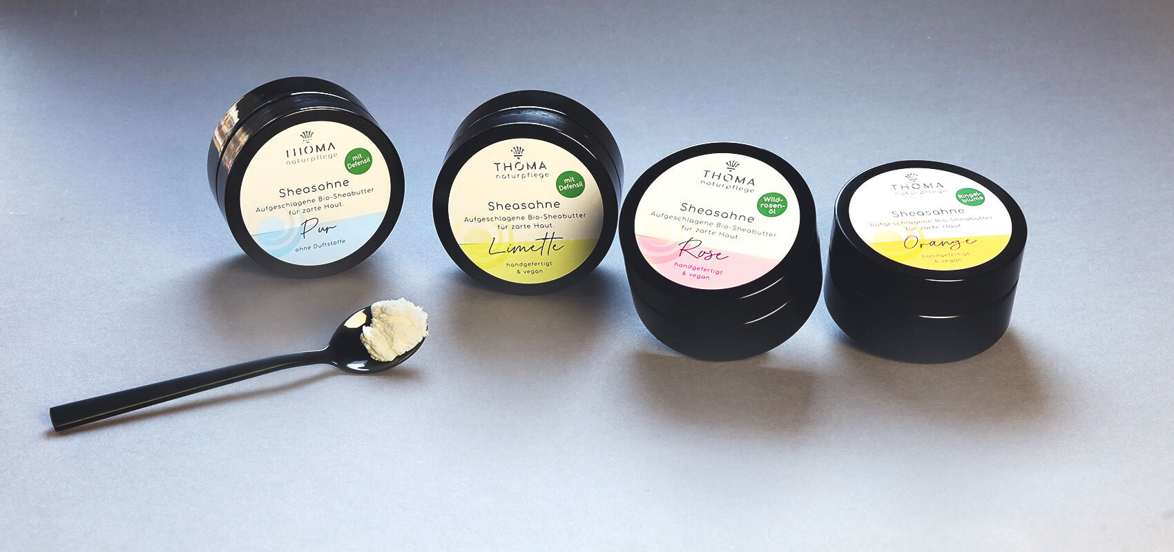 Vier Dosen THOMA Sheasahne in den Sorten Pur, Limette, Rose und Orange, auf grauem Untergrund präsentiert. Ein schwarzer Löffel mit einer Portion der aufgeschlagenen Sheabutter liegt im Vordergrund.
