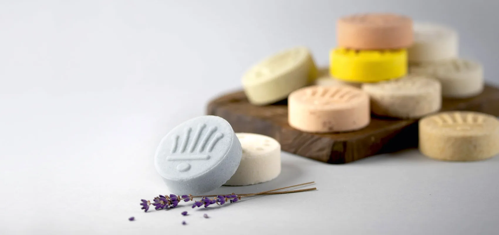 Handgefertigte feste Shampoos in verschiedenen Farben, kunstvoll auf einem Holzbrett und einer hellen Oberfläche arrangiert. Im Vordergrund liegt eine hellblaue Shampoo-Scheibe mit Lavendelzweig.